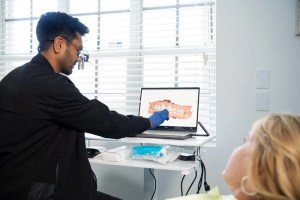 Dentist in black uniform and blue gloves showing digital treatment plan with 3D dental model on laptop to female patient in consultation